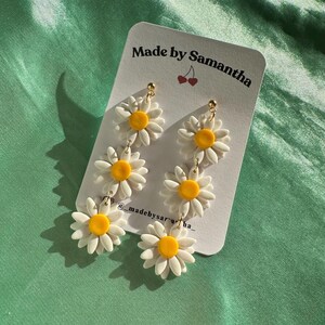 May include: A pair of dangle earrings featuring three white daisy flowers with yellow centers. The earrings are on a white card with the text "Made by Samantha" and "@madebysaracha_" printed on it.