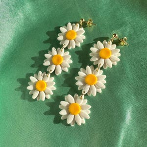 May include: A pair of dangle earrings featuring four white daisy flowers with yellow centers. The earrings are connected by gold chains.