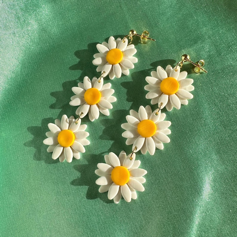 May include: A pair of dangle earrings featuring four white daisy flowers with yellow centers. The earrings are connected by gold chains.
