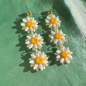 May include: A pair of dangle earrings featuring three white daisy flowers with yellow centers on each earring. The earrings are connected by gold chains.