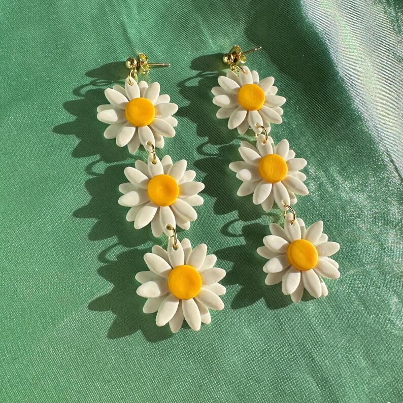 May include: A pair of dangle earrings featuring three white daisy flowers with yellow centers on each earring. The earrings are connected by gold chains.