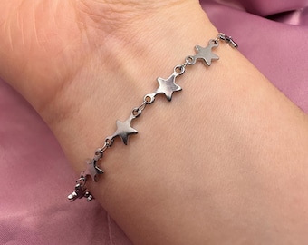 May include: A silver-colored bracelet featuring a series of star-shaped charms. The bracelet is delicate and appears to be made of metal. The stars are evenly spaced along the chain, creating a celestial theme. The bracelet is worn on a wrist.