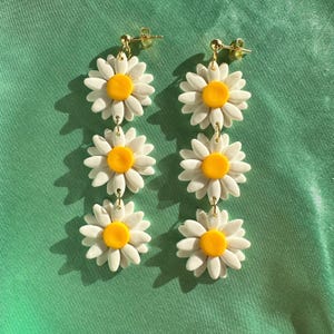 May include: A pair of dangle earrings featuring three white daisy flowers with yellow centers. The earrings are made of polymer clay and have gold-toned hooks.