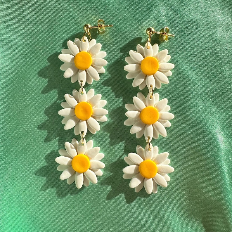 May include: A pair of dangle earrings featuring three white daisy flowers with yellow centers. The earrings are made of polymer clay and have gold-toned hooks.