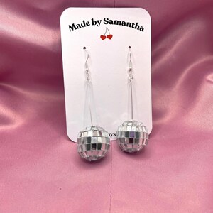May include: A pair of silver disco ball earrings with silver hooks. The earrings are on a white card with the text "Made by Samantha" and a red cherry graphic.