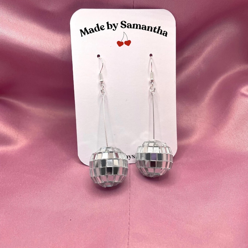 May include: A pair of silver disco ball earrings with silver hooks. The earrings are on a white card with the text "Made by Samantha" and a red cherry graphic.