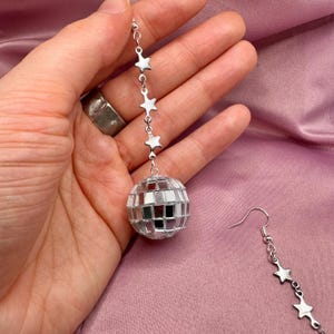 May include: Silver disco ball earrings with a chain of stars hanging from the ball. The earrings are dangling from a person's ear.