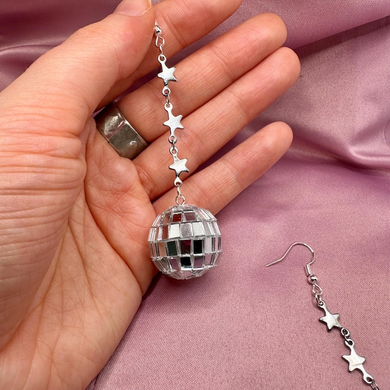 May include: Silver disco ball earrings with a chain of stars hanging from the ball. The earrings are dangling from a person's ear.