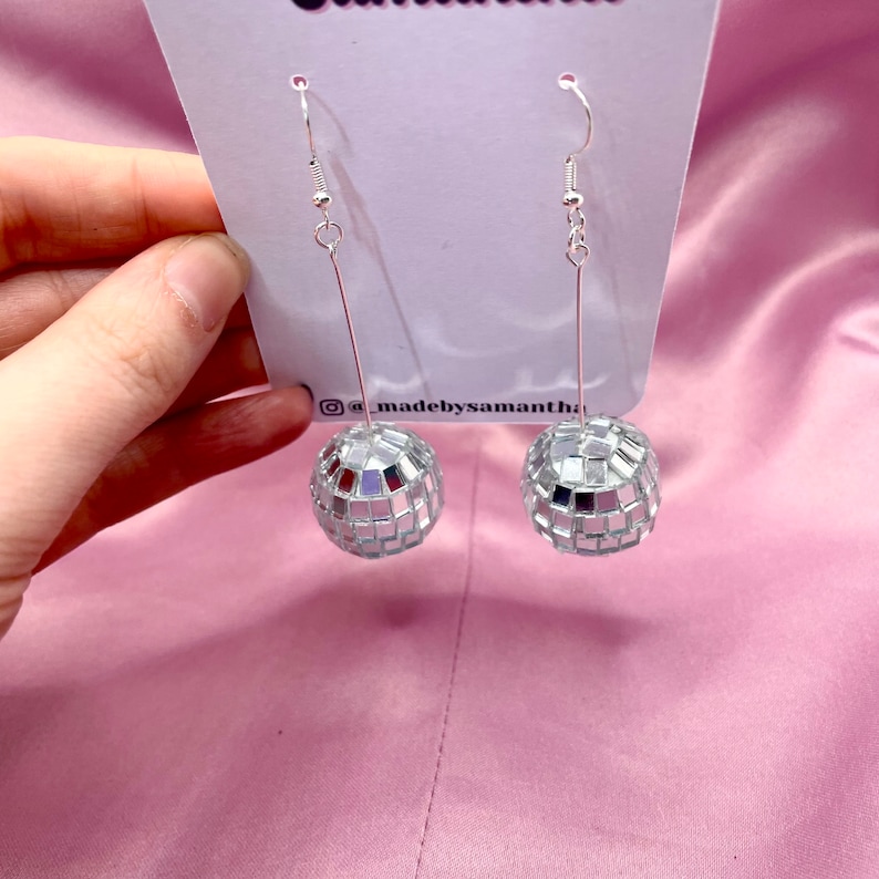 May include: Silver disco ball earrings with a silver hook. The earrings are hanging from a white card with the text "@madebysamantha" on it.
