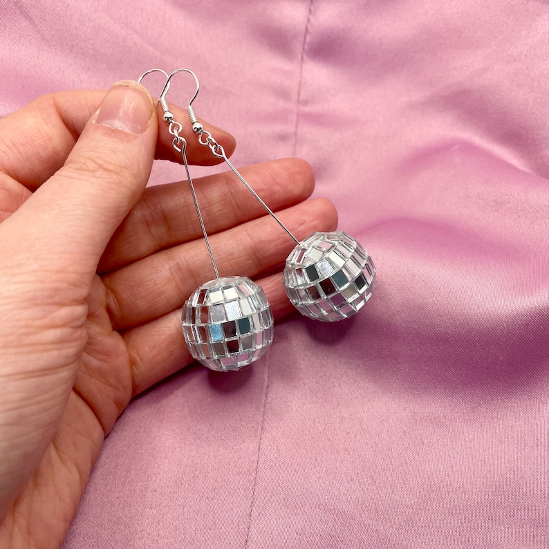 May include: A pair of silver disco ball earrings with a mirrored finish. The earrings are hanging from silver hooks.
