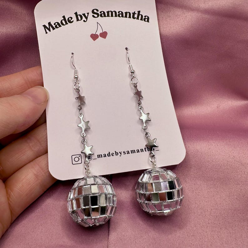 May include: Silver disco ball earrings with a star chain. The earrings are on a white card with the text "Made by Samantha" and a small cherry graphic.