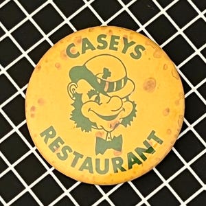 Puede incluir: Un botón redondo y amarillo de Casey's Restaurant con un gráfico de duende verde y texto. El texto dice "CASEYS" en la parte superior y "RESTAURANT" en la parte inferior. El botón está sobre una superficie a cuadros en blanco y negro.