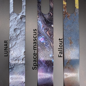 May include: Three rectangular metal keychains with a hole at the top. The keychains have different designs: a lunar surface, a space-themed pattern, and a rusted, weathered appearance. The words "Lunar", "Space-mascus", and "Fallout" are printed on the keychains.
