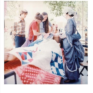 May include: Three people are looking at a quilt with a red, white, and blue pattern. The quilt is spread out on a table. A woman in a long blue dress is standing to the right of the table.