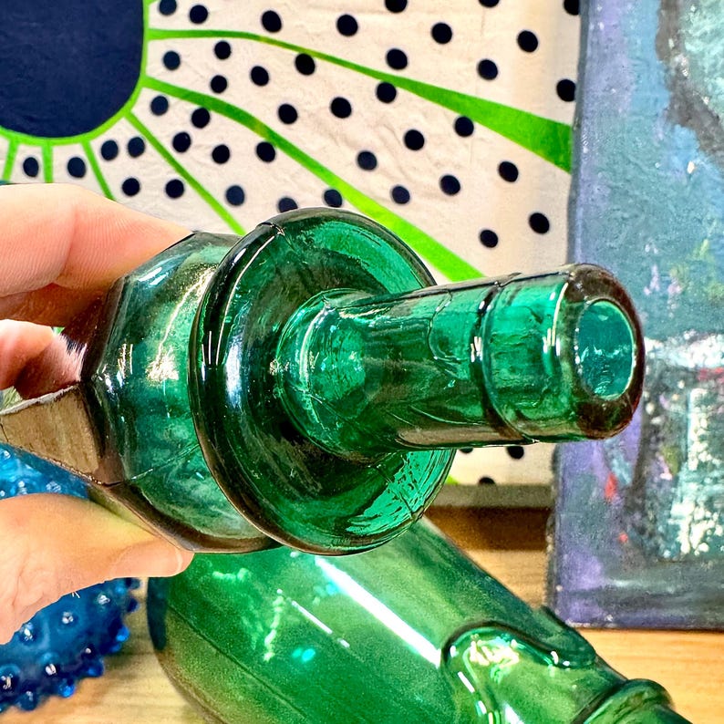 May include: A close-up of a green glass bottle with a round, wide mouth. The bottle is partially obscured by a hand holding it.