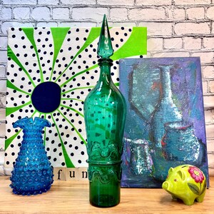 May include: A teal blue glass bottle with a pointed top and a wavy design. The bottle is sitting on a wooden surface in front of a green and white polka dot canvas and a purple abstract painting.