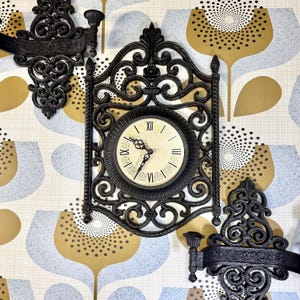 May include: A black metal clock with ornate scrollwork and a white face with black Roman numerals. The clock is mounted on a wall with a floral pattern wallpaper.