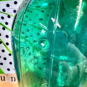 May include: A teal green glass bottle with a textured surface. The bottle has a round body and a narrow neck.