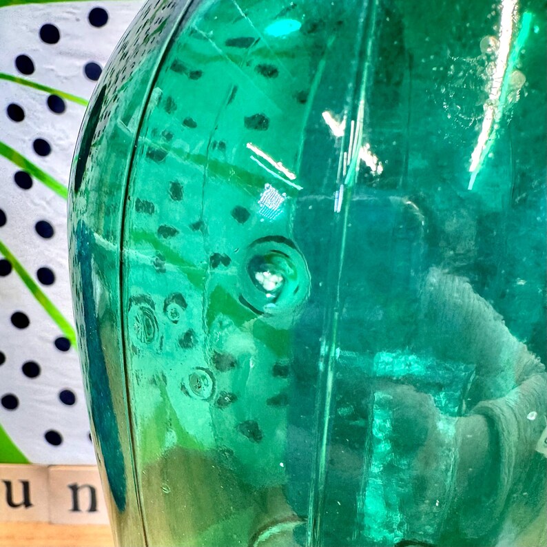 May include: A teal green glass bottle with a textured surface. The bottle has a round body and a narrow neck.