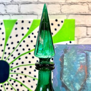 May include: A green glass bottle with a pointed stopper. The bottle is a deep emerald green color and has a textured surface. The stopper is a sharp point and is also green.