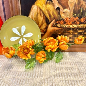 May include: A decorative arrangement featuring a yellow-green plate with a white floral design, complemented by vibrant orange artificial flowers with green leaves. The arrangement is set against a textured, off-white surface and a framed painting.