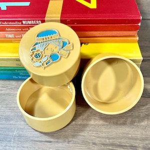 Può includere: Tre contenitori di plastica gialla con coperchi. Il contenitore superiore presenta un'illustrazione colorata di una ragazza con un cappello blu e un fiore.