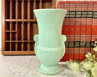 May include: A light green ceramic vase with a speckled finish and two handles. The vase has a classic urn shape and a wide base.