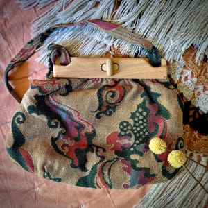 Op de afbeelding: Een vintage handtas met een houten sluiting en handvat. De tas heeft een beige stof met een kleurrijk paisley patroon in tinten rood, groen en paars. Twee gele pompons zijn bij de tas geplaatst.