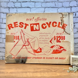 Könnte beinhalten: Eine Vintage-Schachtel für ein Rest 'N Cycle-Trainingsgerät. Die Schachtel ist weiß mit rotem Text und einem Bild einer Frau, die das Gerät benutzt. Der Text auf der Schachtel lautet "new! effective Rest 'N Cycle" und "new! 12,98 $". Die Schachtel sagt auch "entspanntes Trainingsprogramm für die ganze Familie in absoluter Privatsphäre zu Hause" und "lässt sich flach zusammenklappen für einfache Aufbewahrung im Schrank oder Regal".