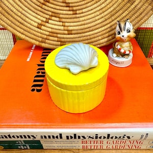 Pode incluir: Uma caixa de bugigangas de cerâmica amarela com uma tampa em forma de concha branca. Uma pequena figura de esquilo com um salva-vidas está por perto. A caixa está em cima de um livro laranja.