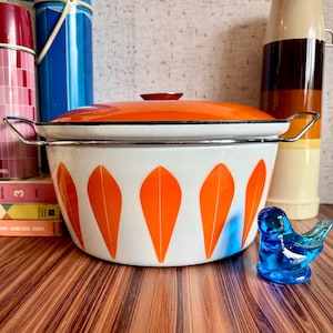 May include: A vintage white enamel pot with an orange lid and handles. The pot features a pattern of orange leaf shapes. A blue glass bird figurine sits to the right of the pot. Other vintage items are in the background.