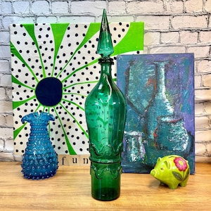 May include: A teal green glass bottle with a pointed top and a wavy design around the base. The bottle is sitting on a wooden surface in front of two paintings. One painting has a green and white polka dot design and the other painting has a blue and purple abstract design.