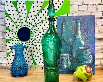 May include: A teal green glass bottle with a pointed top and a wavy design around the base. The bottle is sitting on a wooden surface in front of two paintings. One painting has a green and white polka dot design and the other painting has a blue and purple abstract design.