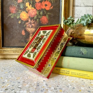 Könnte beinhalten: Eine rechteckige rote Box mit goldenen Verzierungen und einem dekorativen goldenen Verschluss. Die Box hat eine cremefarbene Mittelplatte mit goldenen Akzenten und grünen Blattmotiven. Die Box ist angewinkelt und zeigt ihre verzierten Details.