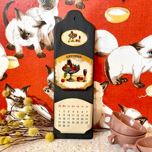 May include: A black calendar with a fruit and wine design on the top section. The calendar has a white background with black lettering and numbers. The calendar is hanging on a wall with a red and white floral pattern.