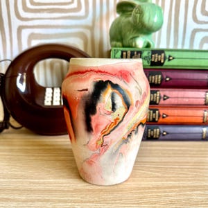 May include: A ceramic vase with a unique abstract design. The vase features a blend of pink, black, and gold colors. The vase is cylindrical with a slightly flared top. The background includes a vintage telephone and a stack of books.