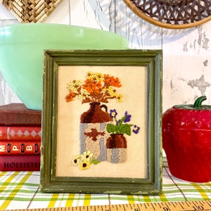 May include: A framed embroidery of a floral arrangement in two brown ceramic vases. The flowers are yellow, orange, and purple. The frame is green.