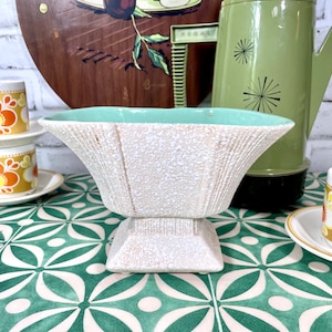 Op de afbeelding: Een witte keramische plantenpot met een groene rand en een getextureerd oppervlak. De pot heeft een vierkante basis en een brede, afgeronde bovenkant. Hij staat op een groene en witte betegelde ondergrond.