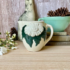 Könnte beinhalten: Eine weiße Keramikkanne mit grünem Blumendesign. Die Kanne hat einen Henkel und eine breite Öffnung. Das Blumendesign zeigt eine große weiße Blume mit grünen Blättern.