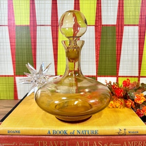 May include: Amber glass decanter with a round stopper. The decanter has a wide base and a narrow neck. The stopper is clear glass and has a round top.