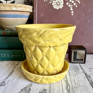 Op de afbeelding: Een felgele keramische plantenpot met bijpassende schotel. De pot is voorzien van een verhoogd ruitpatroon op de romp en een decoratief bladmotief rond de rand. De schotel heeft ook een bijpassend bladontwerp langs de rand.