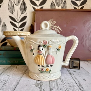 May include: Cream-colored ceramic teapot with a lid and handle. The teapot features a raised relief design of two figures holding hands, surrounded by flowers. The teapot is on a wooden surface.