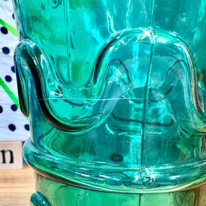 May include: A teal blue glass bottle with a unique, wavy design. The bottle has a wide mouth and a rounded base.