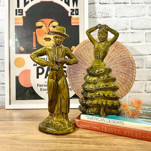 May include: Two ceramic figurines, a man and a woman, dressed in traditional Spanish clothing. The man is holding a guitar and the woman is wearing a ruffled dress. The figurines are green and brown and are standing on a wooden surface.