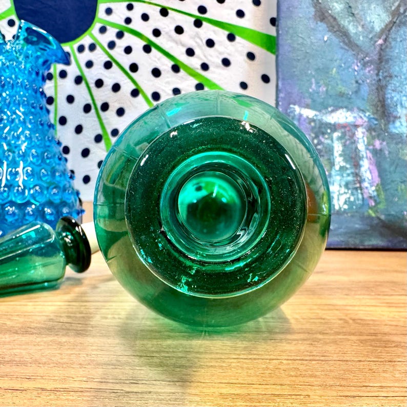 May include: A teal green glass bottle with a round bottom and a wide opening. The bottle is sitting on a wooden surface.