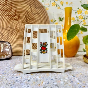 May include: White plastic napkin holder with a red tomato and green leaves design. The holder has a grid pattern and a curved base.