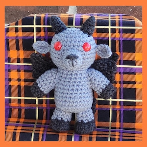 May include: A crocheted plush toy in the shape of a goat-like creature. The toy is primarily light blue with black accents on the horns, wings, and hooves. It has red eyes and is sitting on a plaid patterned surface with orange, purple, and black colors.