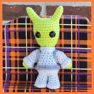 May include: A handmade crochet doll with a green head, large black eyes, and pointed ears. The doll wears a light blue outfit with a turtleneck and matching pants. The doll is sitting on a plaid fabric with orange, purple, and black squares.