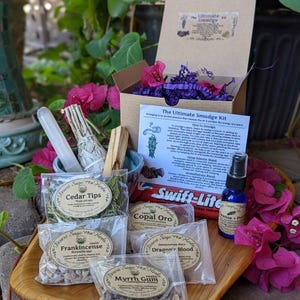 May include: An assortment of spiritual items, including a kit labeled "The Ultimate Smudge Kit," cedar tips, frankincense, copal oro, dragon's blood, and myrrh gum. A blue spray bottle and a white crystal are also visible. The items are arranged on a wooden tray.