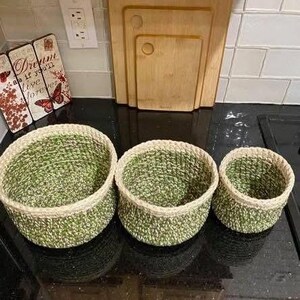 May include: Set of three handmade crochet baskets in varying sizes. The baskets feature a green and white speckled pattern with a natural-colored rim. They are arranged on a black surface, showcasing their woven texture and design.